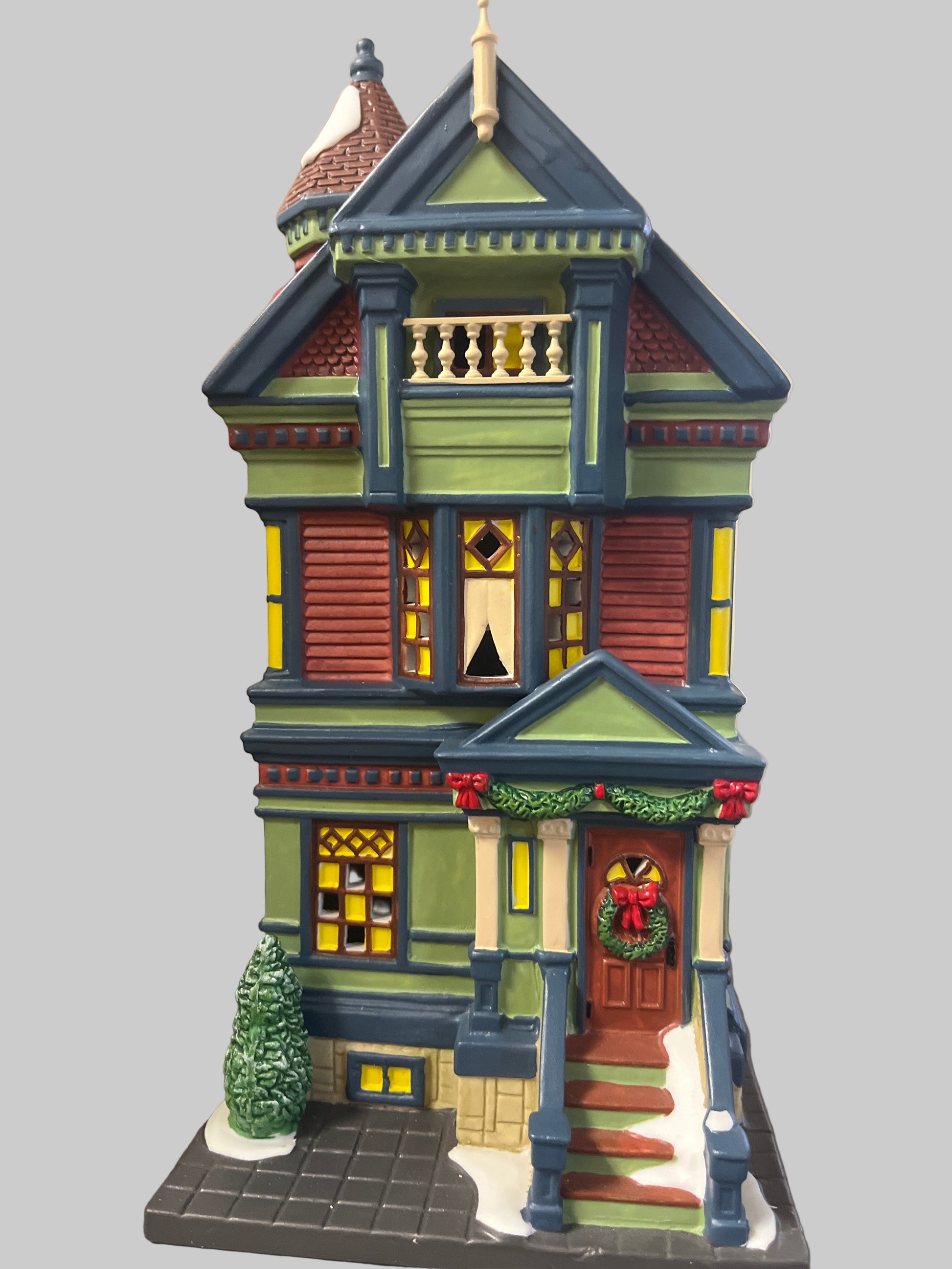 A decorative seasonal village set featuring a green, red, and blue Victorian-style house with a Christmas tree and holiday decorations on the porch.